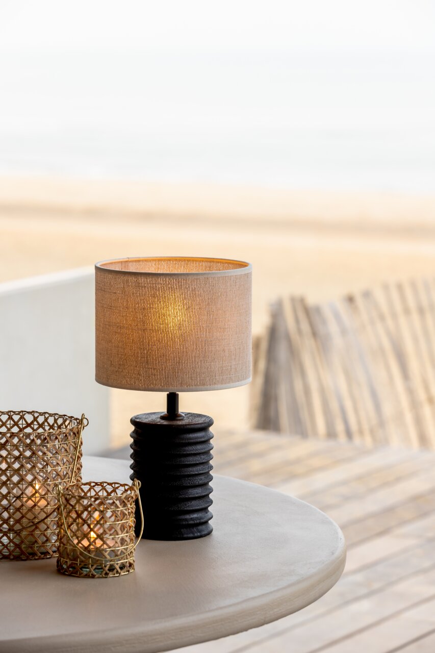 Tischlampe mit einem strukturierten schwarzen Sockel und einem beigen Lampenschirm, auf einem Tisch mit Strandblick im Hintergrund, aus seitlicher Perspektive.