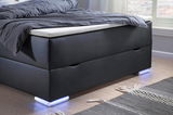 Boxspringbett in seitlicher Perspektive mit dunklem Bezug und leuchtenden Füßen auf Holzboden.