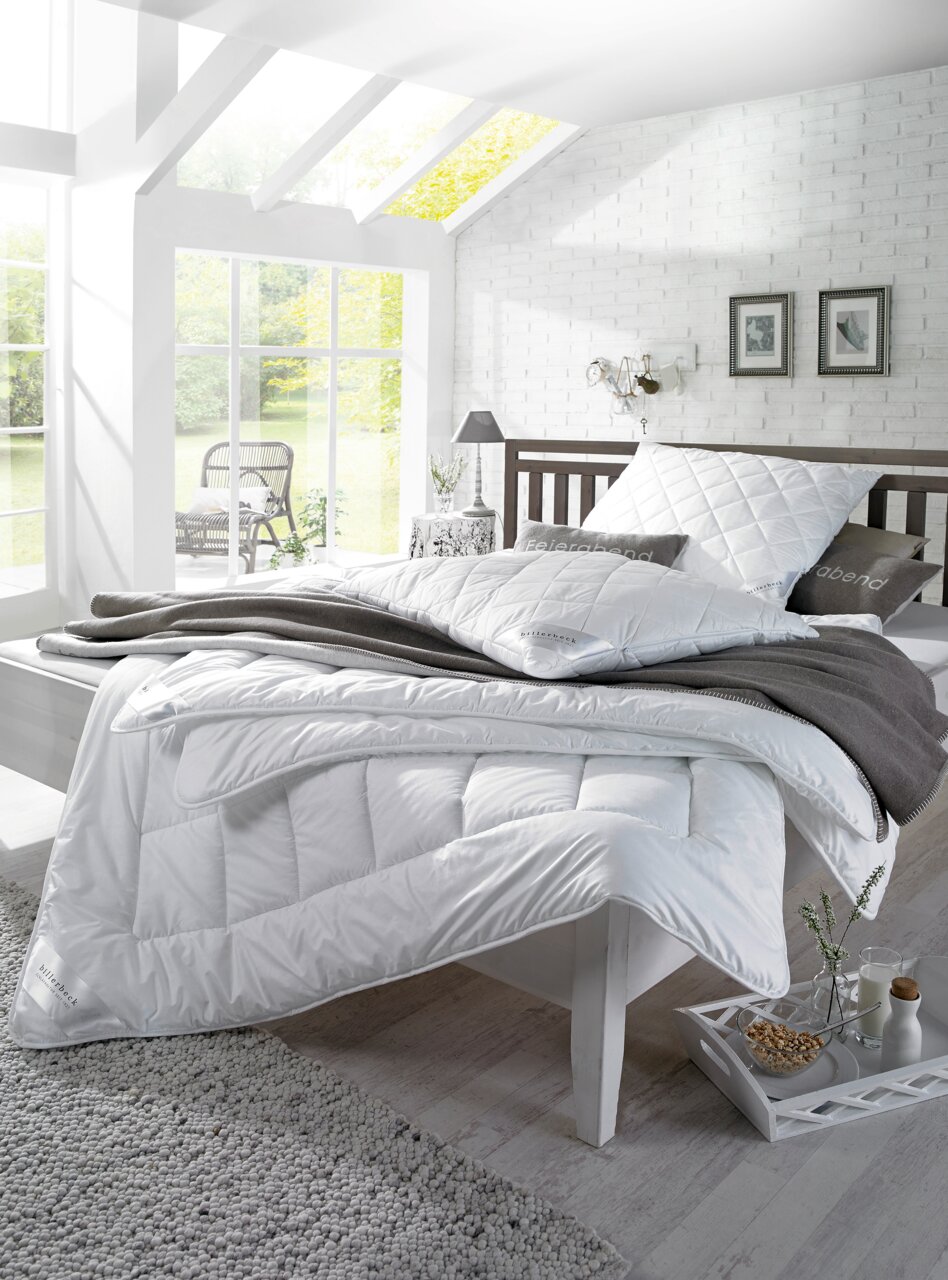 Ein gemütliches Schlafzimmer mit einem weißen, gesteppten Kopfkissen und einer passenden Bettdecke auf einem modernen Bett. Die Perspektive zeigt das Bett von der Seite, mit einem Fenster im Hintergrund, das natürliches Licht hereinlässt.