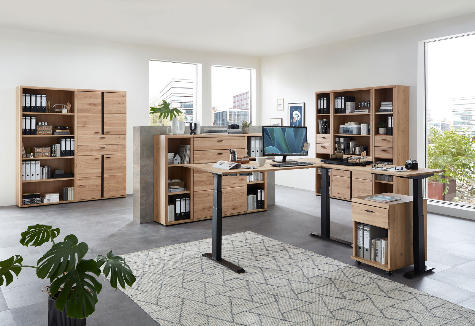 Moderne Büromöbel in einem hellen Raum mit großen Fenstern, bestehend aus einem höhenverstellbaren Eckschreibtisch aus Melamin, umgeben von Aktenschränken und Regalen aus Holz. Perspektive: Frontalansicht.