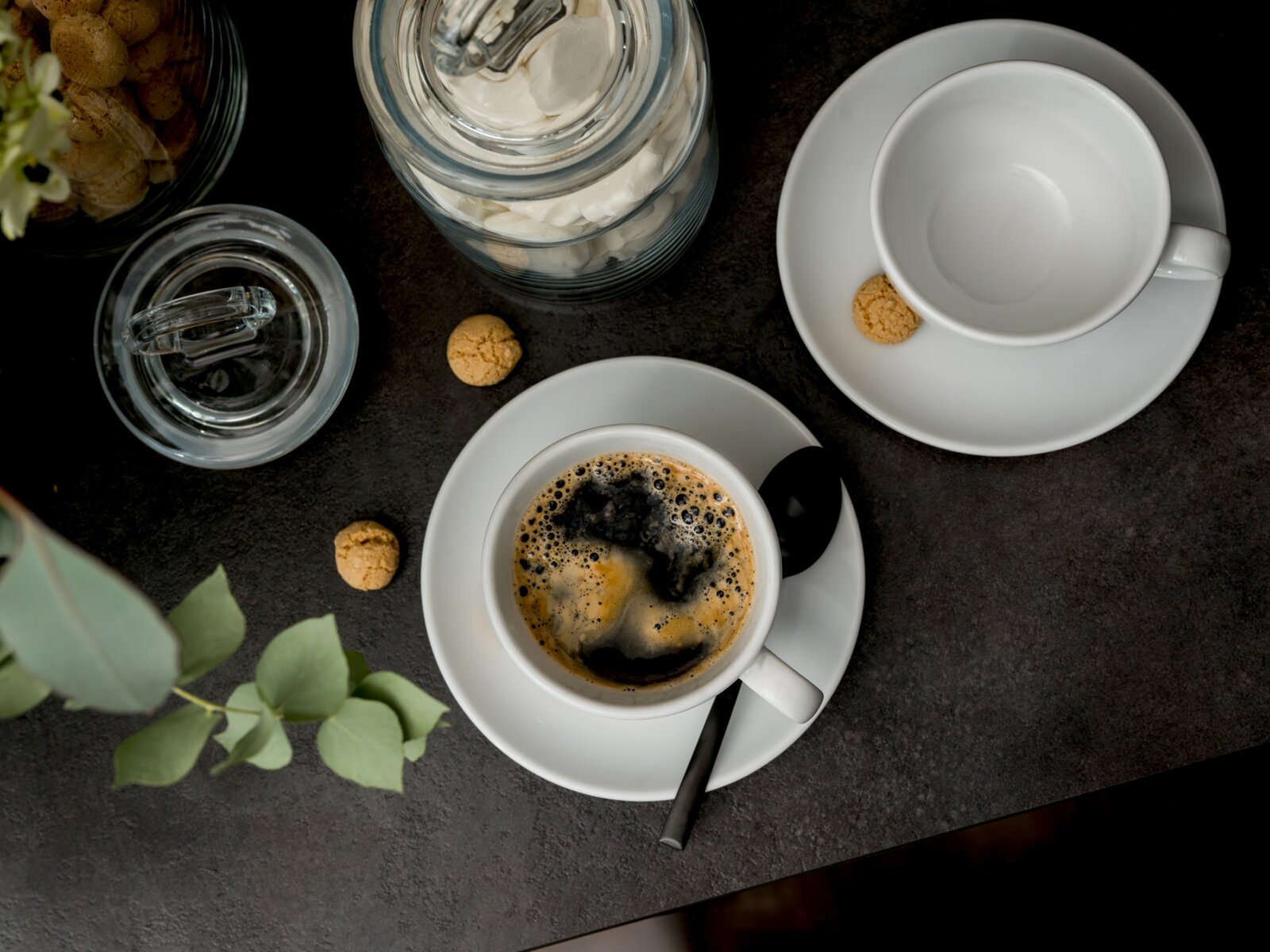 Draufsicht auf eine weiße Kaffeetasse mit schwarzem Kaffee und Löffel auf Untertasse, daneben eine leere Tasse, ein Glas mit Baiser und kleine Kekse auf dunkler Tischplatte