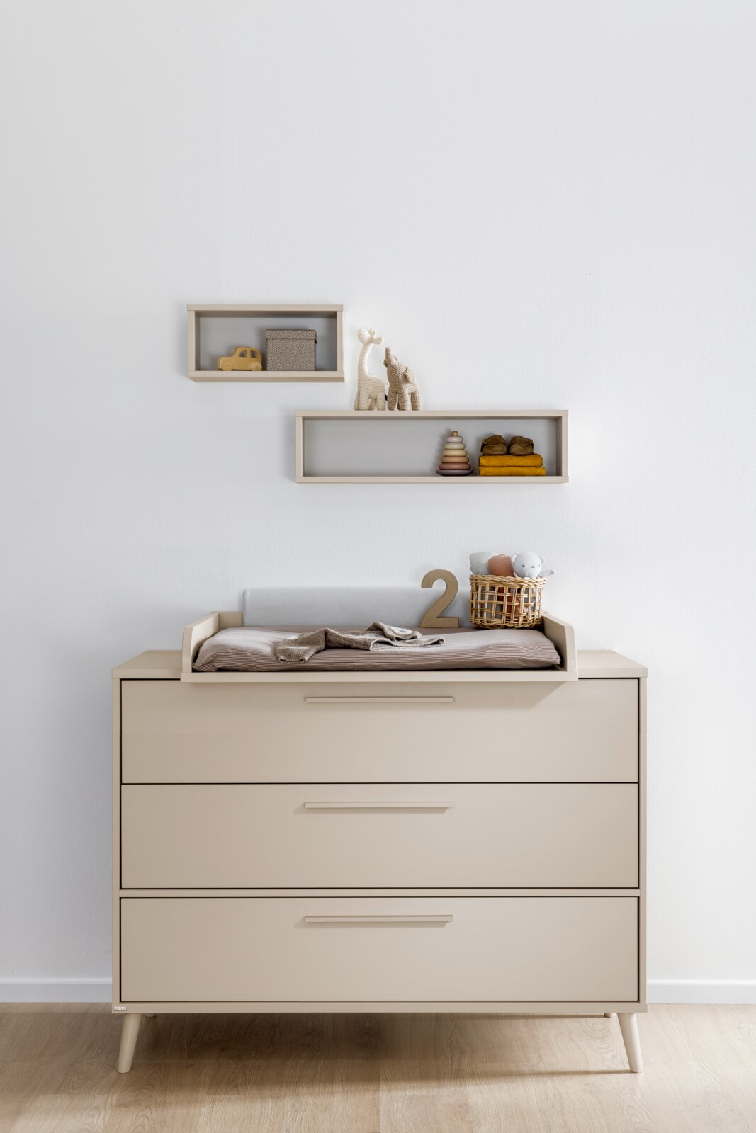 Beige Wickelkommode mit drei Schubladen und Wickelaufsatz im Kinderzimmer, frontal aus der Vorderansicht fotografiert; auf dem Aufsatz liegen Wickelunterlage, Decke, Korb und Deko, darüber zwei schwebende Wandregale.