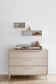 Beige Wickelkommode mit drei Schubladen und Wickelaufsatz im Kinderzimmer, frontal aus der Vorderansicht fotografiert; auf dem Aufsatz liegen Wickelunterlage, Decke, Korb und Deko, darüber zwei schwebende Wandregale.