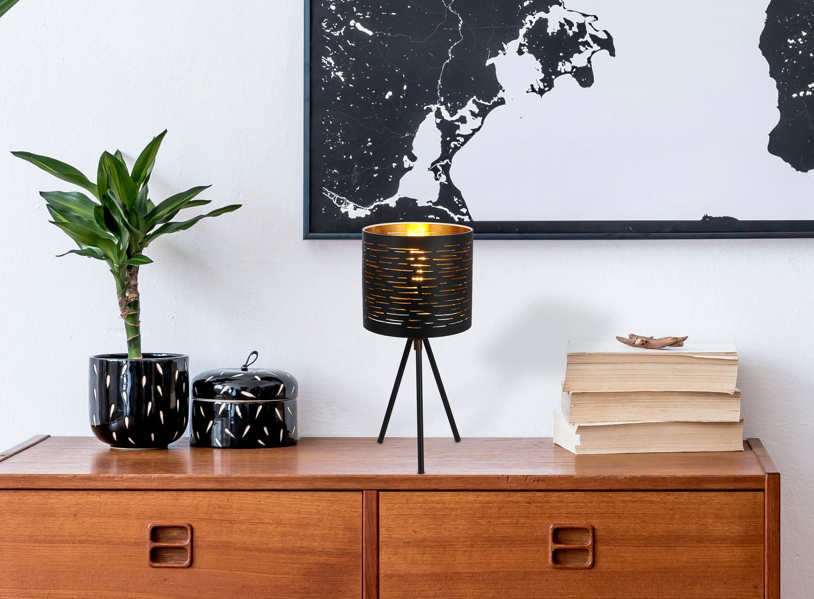 Moderne Tischleuchte mit schwarzem, perforiertem Lampenschirm auf dreibeinigem Ständer, auf einem Holztisch neben einer Pflanze und Büchern, aus seitlicher Perspektive.