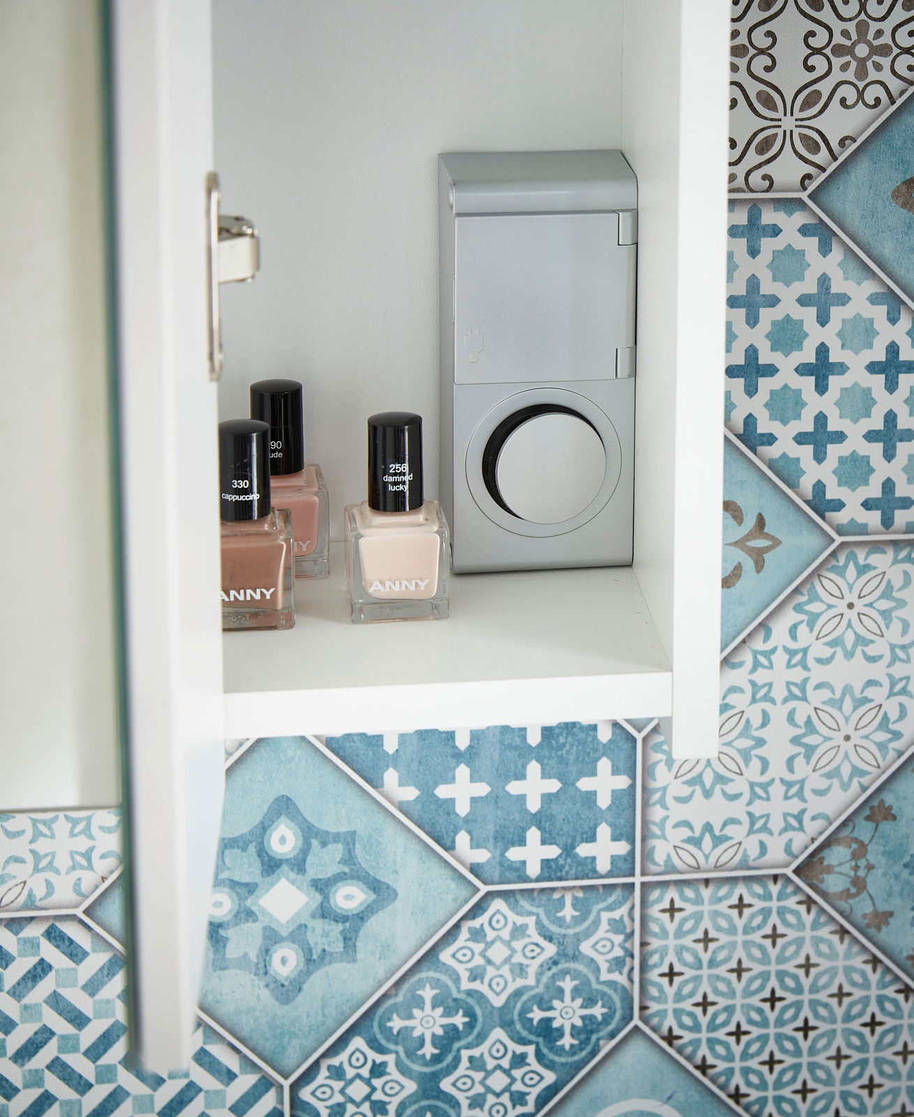 Innenansicht eines Spiegelschranks mit Nagellackflaschen und einer Steckdose, umgeben von dekorativen blauen Fliesen.