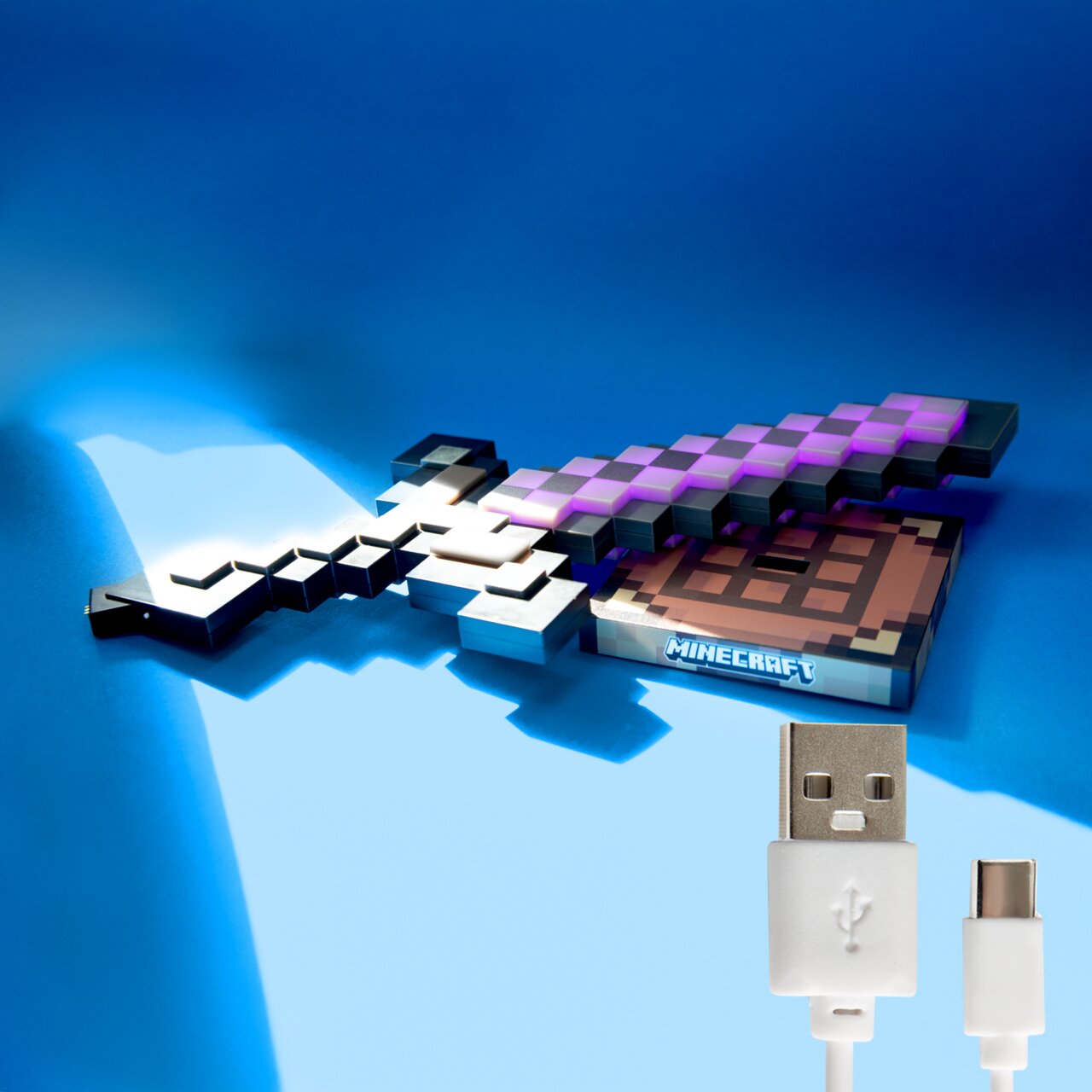 Minecraft Pixel-Schwert als leuchtendes Deko- oder Spielzeug-Accessoire auf blauem Hintergrund, schräg von oben fotografiert; unten rechts ein weißes USB-A-auf-USB-C-Ladekabel im Detail.