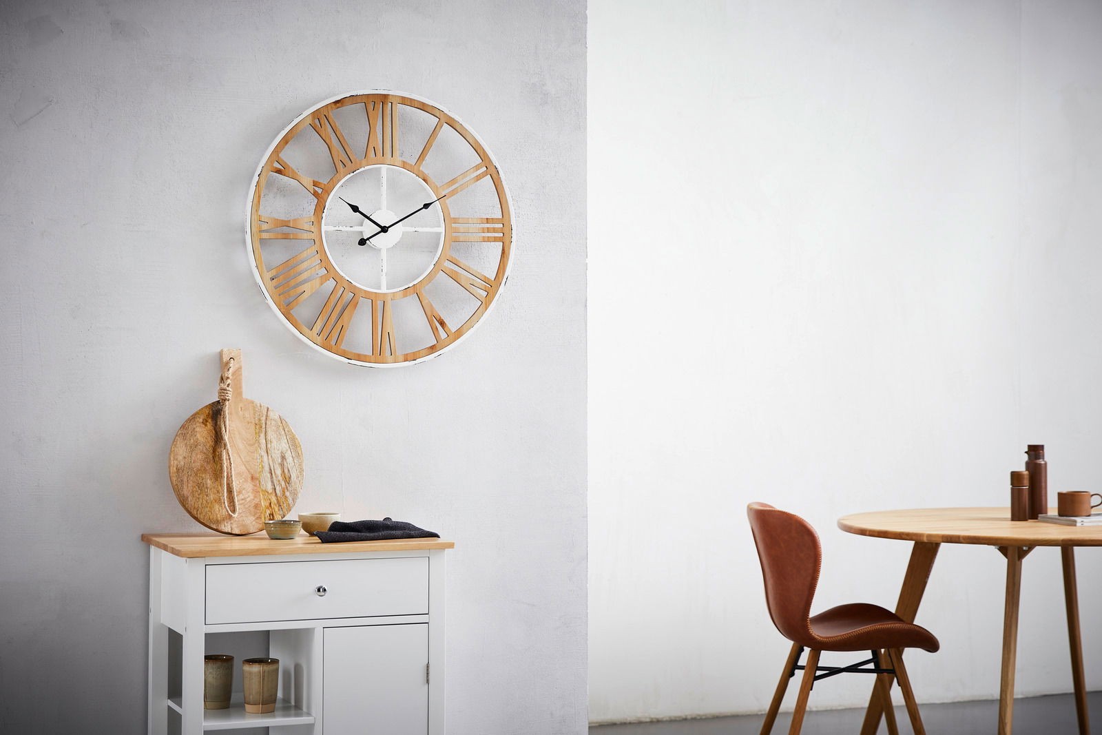 Große Wanduhr mit römischen Ziffern in Holzoptik, an einer hellen Wand hängend, im modernen Wohnambiente mit Tisch und Stuhl. Frontalansicht.