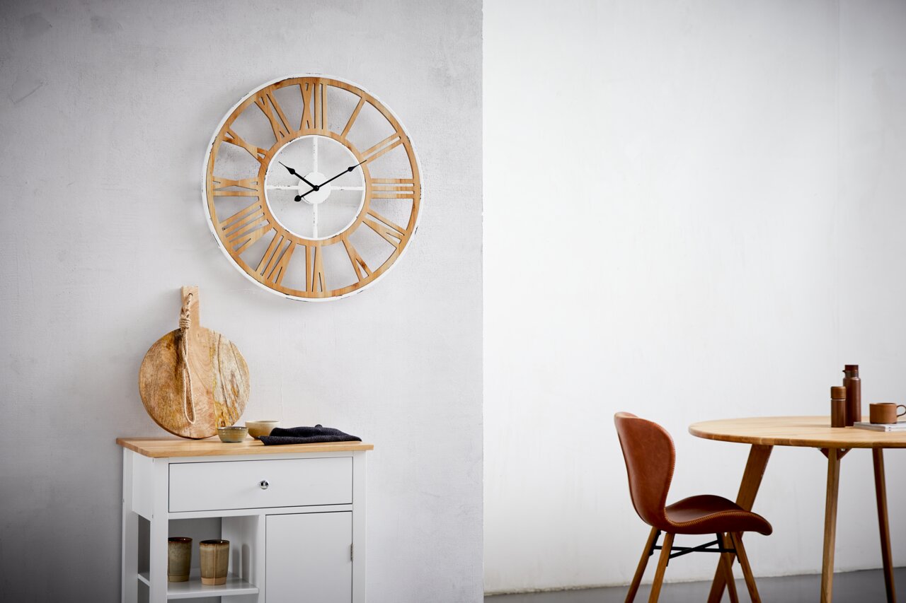 Grote wandklok met Romeinse cijfers in houtlook, hangend aan een lichte muur, in een moderne woonkamer met tafel en stoel. Vooraanzicht.