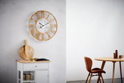 Grote wandklok met Romeinse cijfers in houtlook, hangend aan een lichte muur, in een moderne woonkamer met tafel en stoel. Vooraanzicht.