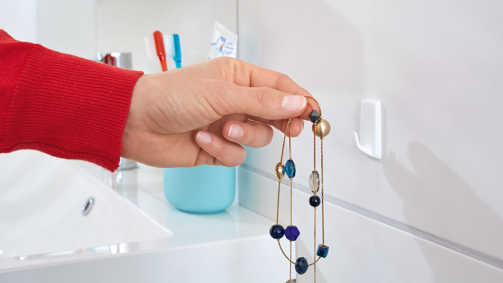 Nahaufnahme eines weißen tesa Powerstrips Hakens an einer Badezimmerwand, an dem eine Hand eine bunte Halskette aufhängt.