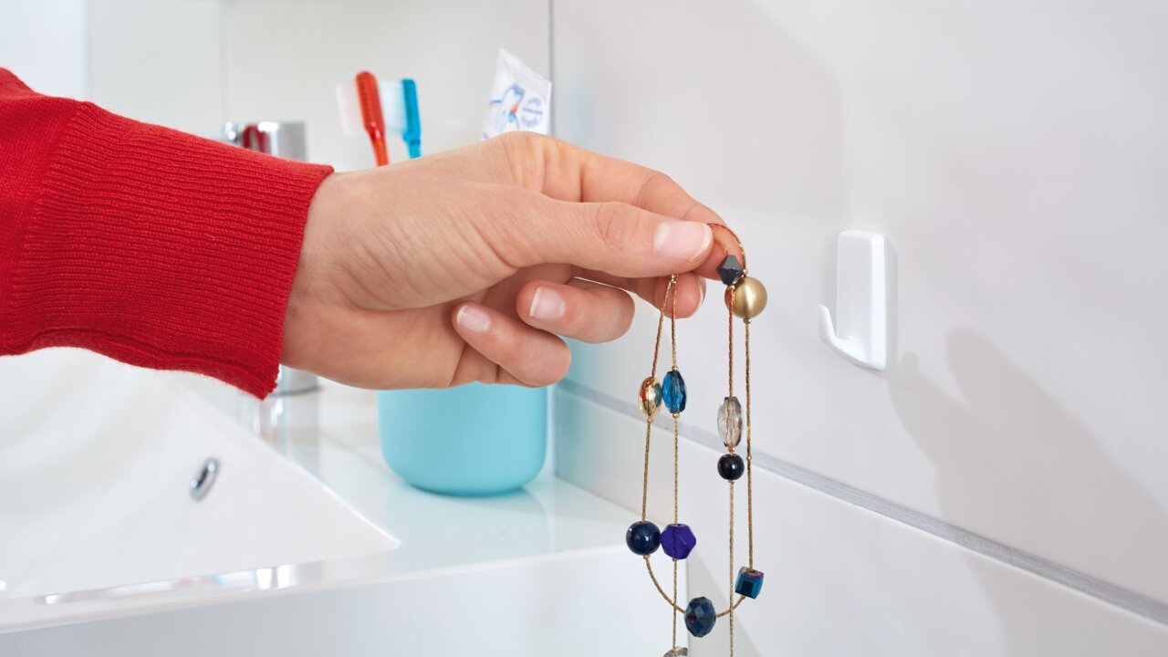 Tesa Powerstrips Haken 3-tlg. Nahaufnahme eines weißen tesa Powerstrips Hakens an einer Badezimmerwand, an dem eine Hand eine bunte Halskette aufhängt.