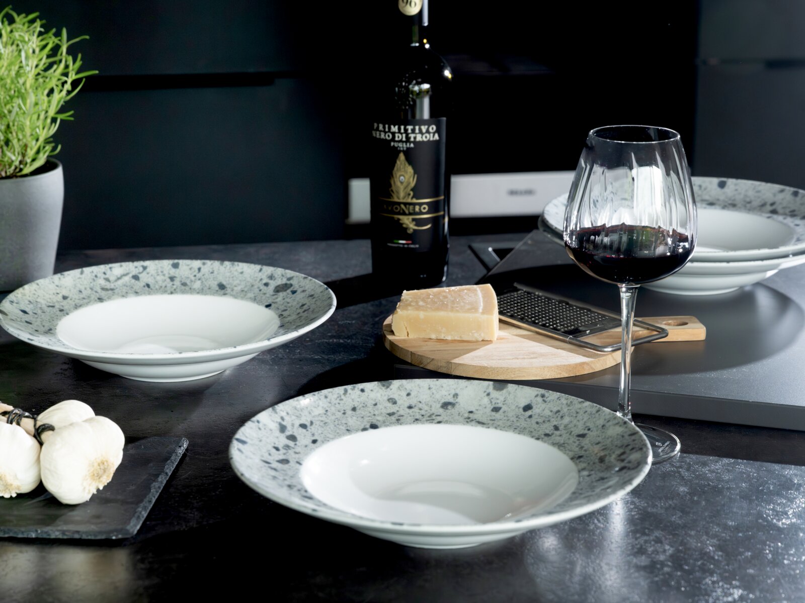 Schwarz-weiß gesprenkelte Suppenteller auf dunklem Tisch mit Rotweinglas, Weinflasche und Parmesan auf Holzbrett; Perspektive: leicht erhöhte Tischansicht (3/4-Ansicht).