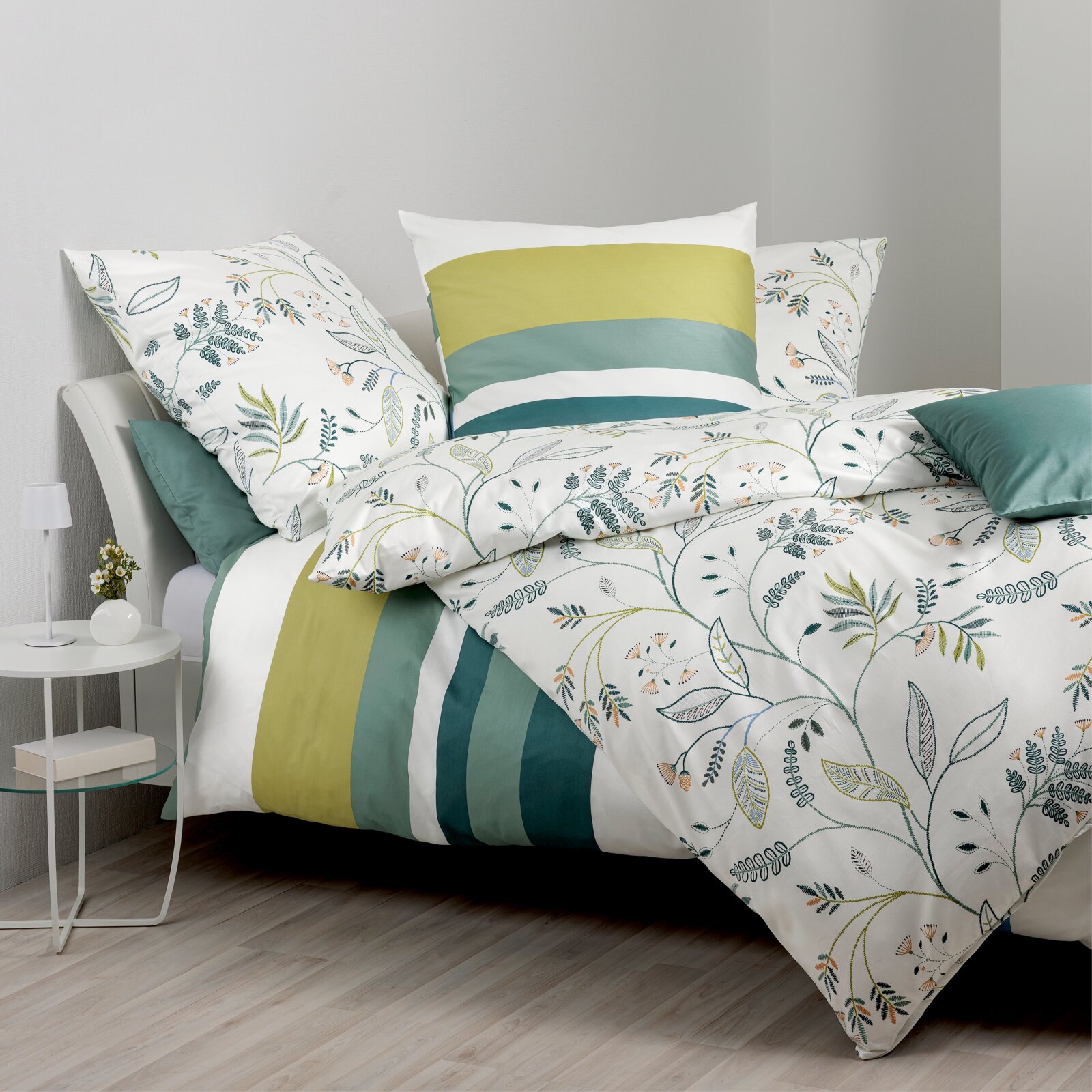 Bettwäsche-Set mit botanischem Blätter- und Zweigmuster in Grün- und Naturtönen, kombiniert mit breiten Streifen in Olivgrün, Mint und Petrol; auf einem Bett arrangiert, fotografiert in schräger Frontansicht.