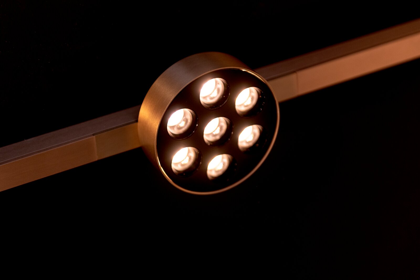Ronde messingkleurige LED-spot met zeven lenzen op een rail, warm wit licht, gefotografeerd in schuin bovenaanzicht tegen een donkere achtergrond