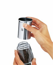 Nahaufnahme einer Gewürzmühle De Luxe aus Edelstahl, die von zwei Händen gehalten wird. Die obere Hälfte wird abgenommen, um die Pfefferkörner im Glasbehälter darunter zu zeigen.