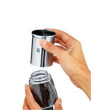 Nahaufnahme einer Gewürzmühle De Luxe aus Edelstahl, die von zwei Händen gehalten wird. Die obere Hälfte wird abgenommen, um die Pfefferkörner im Glasbehälter darunter zu zeigen.