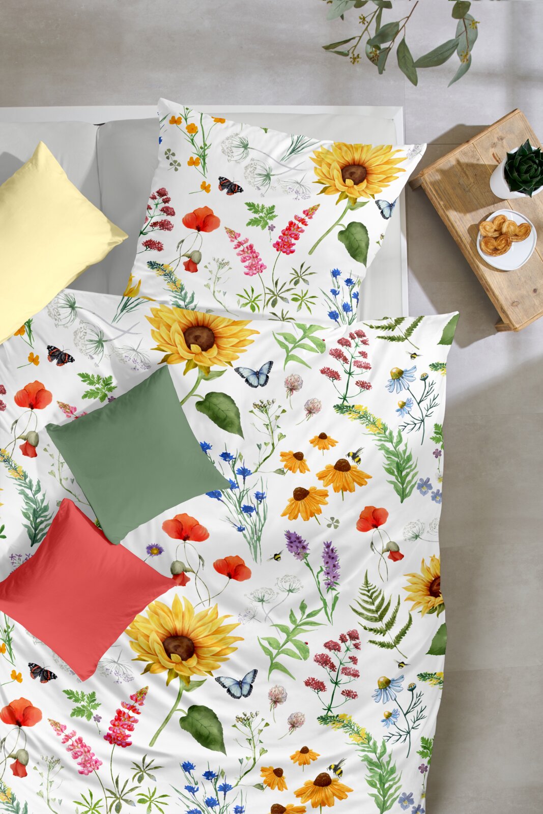 Draufsicht auf ein Bett mit weißer Bettwäsche im bunten Blumen- und Schmetterlingsmuster (Sonnenblumen, Mohn, Wiesenblumen) sowie farbigen Zierkissen; daneben ein Holzhocker mit Pflanze und Gebäck.