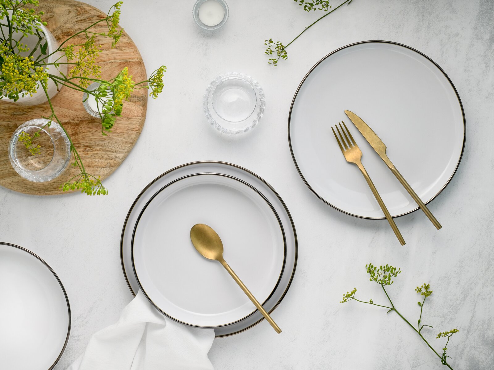 Draufsicht auf ein minimalistisches Tischgedeck mit weißen Tellern mit dunklem Rand, goldfarbenem Besteck und kleinen Glas-Schälchen auf heller Steinoptik, dekoriert mit grünen Blütenzweigen und Holztablett.