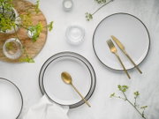 Draufsicht auf ein minimalistisches Tischgedeck mit weißen Tellern mit dunklem Rand, goldfarbenem Besteck und kleinen Glas-Schälchen auf heller Steinoptik, dekoriert mit grünen Blütenzweigen und Holztablett.