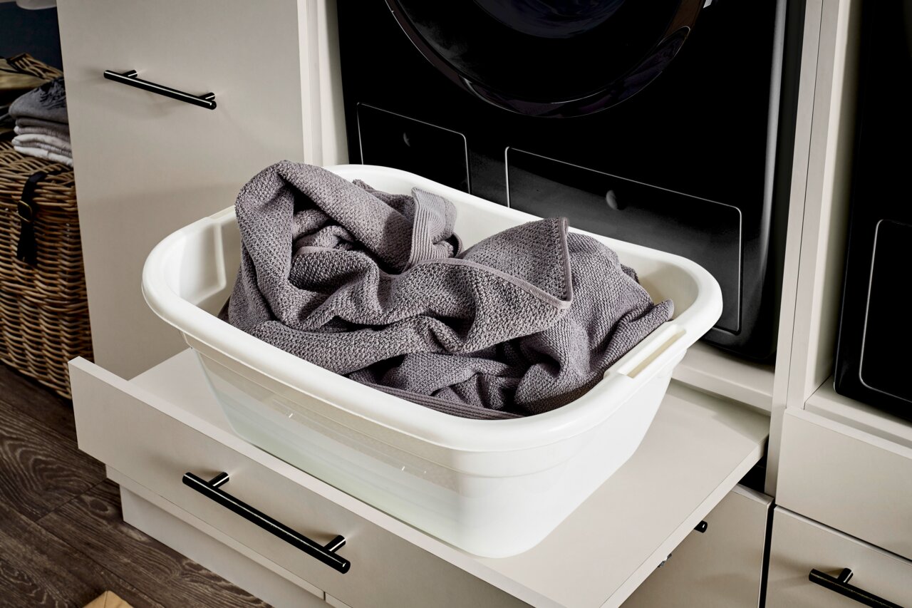 Weiße Kunststoff-Wäschewanne mit grauen Handtüchern auf ausgezogenem Schubladenauszug vor schwarzer Frontlader-Waschmaschine, Perspektive: leicht von oben und schräg von vorn