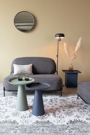 Zwei moderne Beistelltische in verschiedenen Farben, im Vordergrund einer stilvollen Wohnzimmerumgebung mit Sofa, Stehlampe und dekorativem Teppich, aus seitlicher Perspektive.