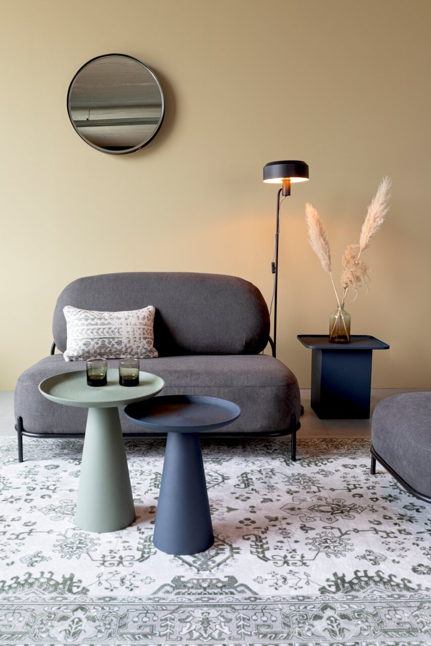 Zwei moderne Beistelltische in verschiedenen Farben, im Vordergrund einer stilvollen Wohnzimmerumgebung mit Sofa, Stehlampe und dekorativem Teppich, aus seitlicher Perspektive.