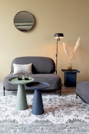 Zwei moderne Beistelltische in verschiedenen Farben, im Vordergrund einer stilvollen Wohnzimmerumgebung mit Sofa, Stehlampe und dekorativem Teppich, aus seitlicher Perspektive.