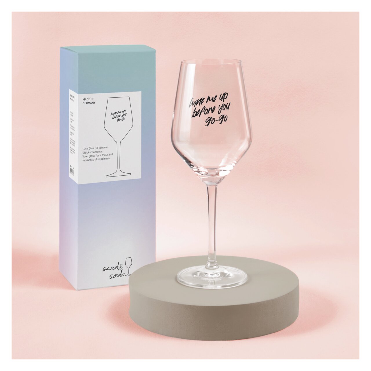 Weinglas mit der Aufschrift 'Wine me up before you go-go', präsentiert auf einem runden Sockel vor einer Verpackung, seitliche Perspektive.