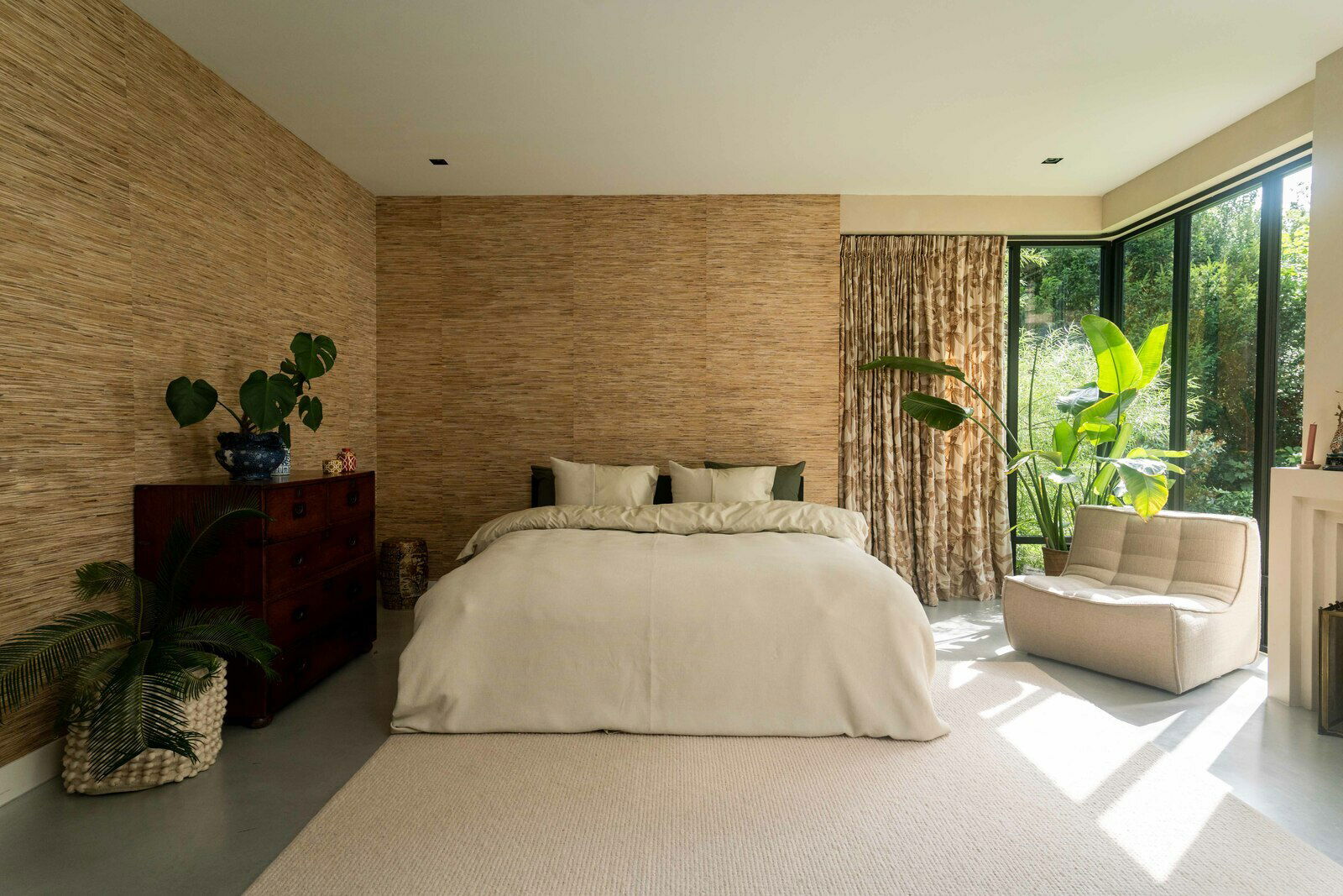 Gemütliches Schlafzimmer mit einem großen Bett in cremefarbener Bettwäsche, umgeben von Pflanzen und natürlichem Licht, aus der Perspektive von vorne.