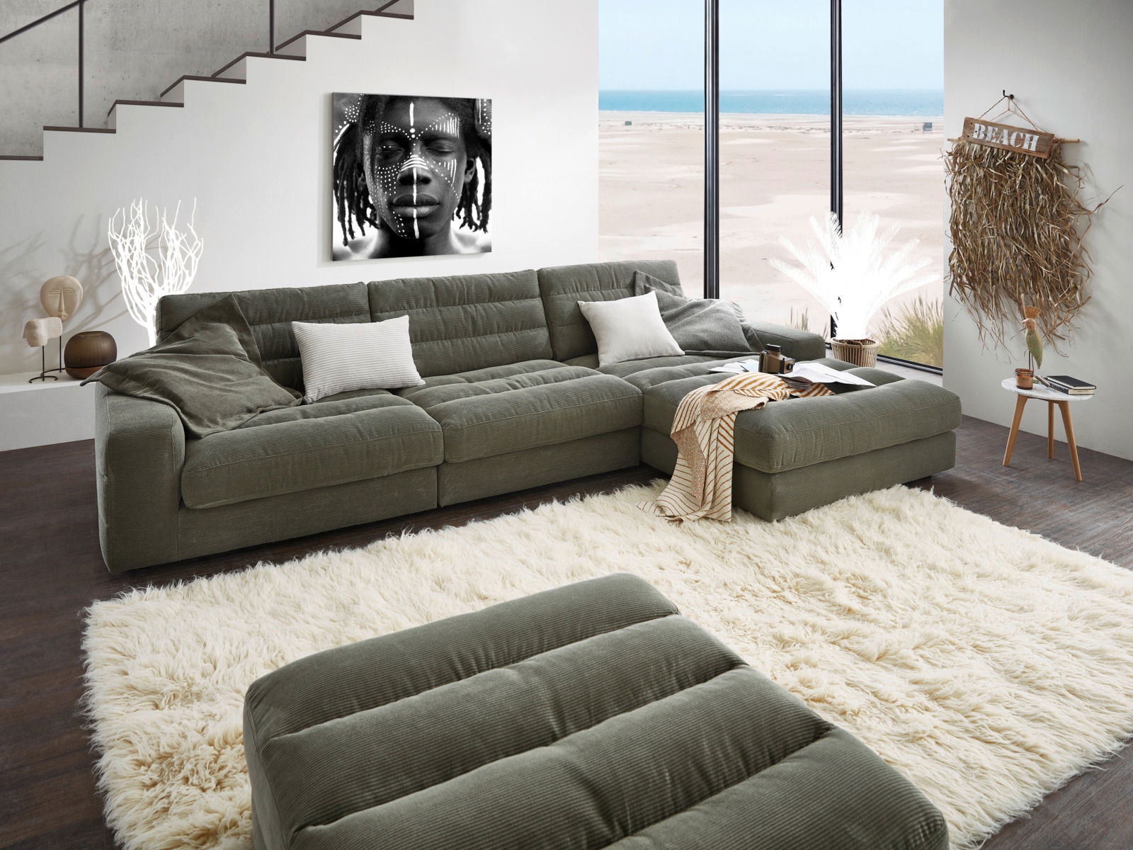 Grüne Longchairkombination in modernem Wohnzimmer mit Blick auf das Meer durch große Fenster, dekoriert mit Kissen und Decke, fotografiert aus einer leicht erhöhten Perspektive.
