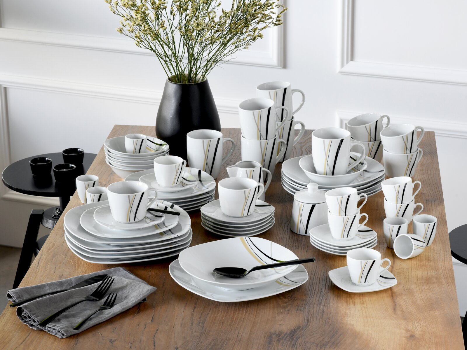 Creatable Kombiservice 62-tlg SQUARE Modernes weißes Porzellan-Geschirrset mit schwarzen und goldenen Linien (Teller, Schalen, Tassen, Becher, Kanne/Zuckerdose) auf Holztisch arrangiert, perspektivisch in leichter Schrägansicht von vorne oben.