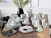 Modernes weißes Porzellan-Geschirrset mit schwarzen und goldenen Linien (Teller, Schalen, Tassen, Becher, Kanne/Zuckerdose) auf Holztisch arrangiert, perspektivisch in leichter Schrägansicht von vorne oben.