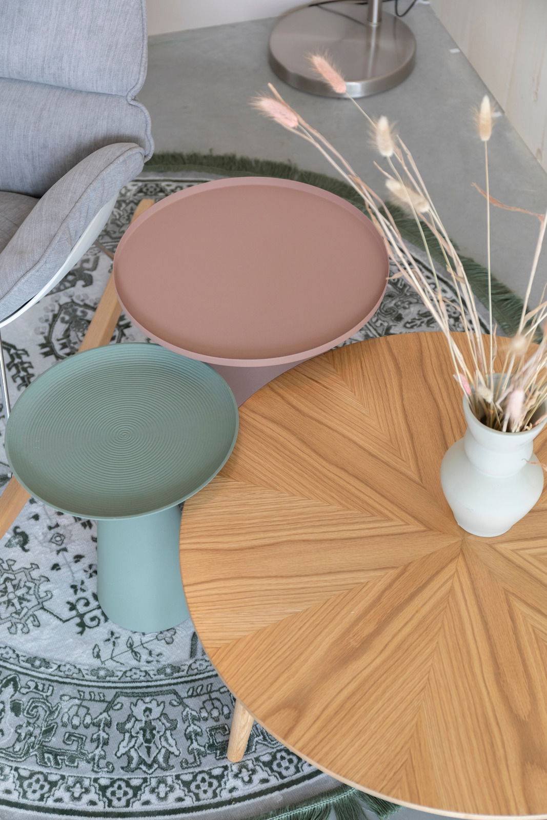 Drei Beistelltische in verschiedenen Farben und Materialien aus der Vogelperspektive: ein runder Holztisch mit Vase, ein grüner Tisch mit geriffelter Oberfläche und ein rosa Tisch auf einem gemusterten Teppich.