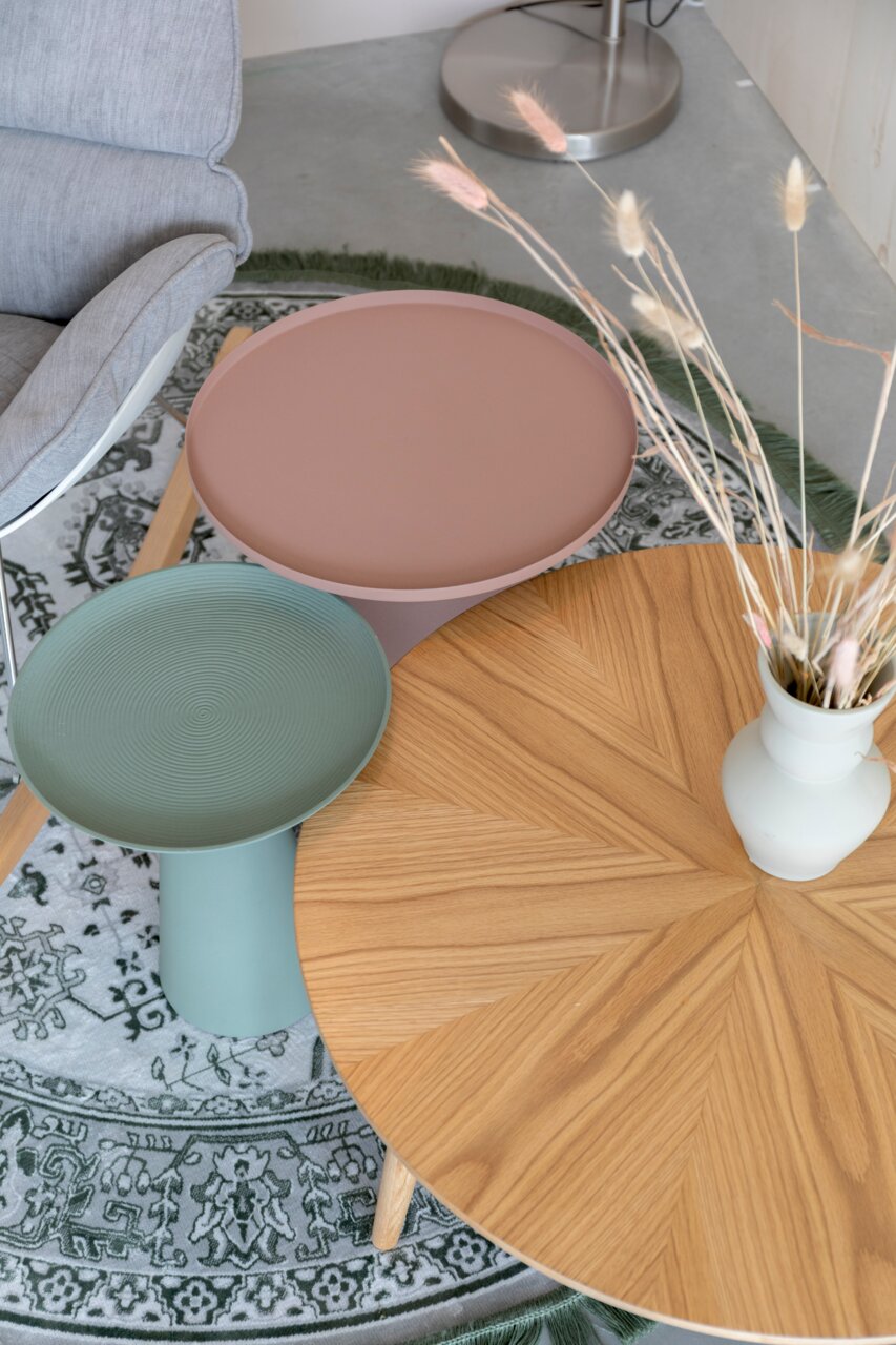 Drei Beistelltische in verschiedenen Farben und Materialien aus der Vogelperspektive: ein runder Holztisch mit Vase, ein grüner Tisch mit geriffelter Oberfläche und ein rosa Tisch auf einem gemusterten Teppich.
