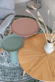 Drei Beistelltische in verschiedenen Farben und Materialien aus der Vogelperspektive: ein runder Holztisch mit Vase, ein grüner Tisch mit geriffelter Oberfläche und ein rosa Tisch auf einem gemusterten Teppich.