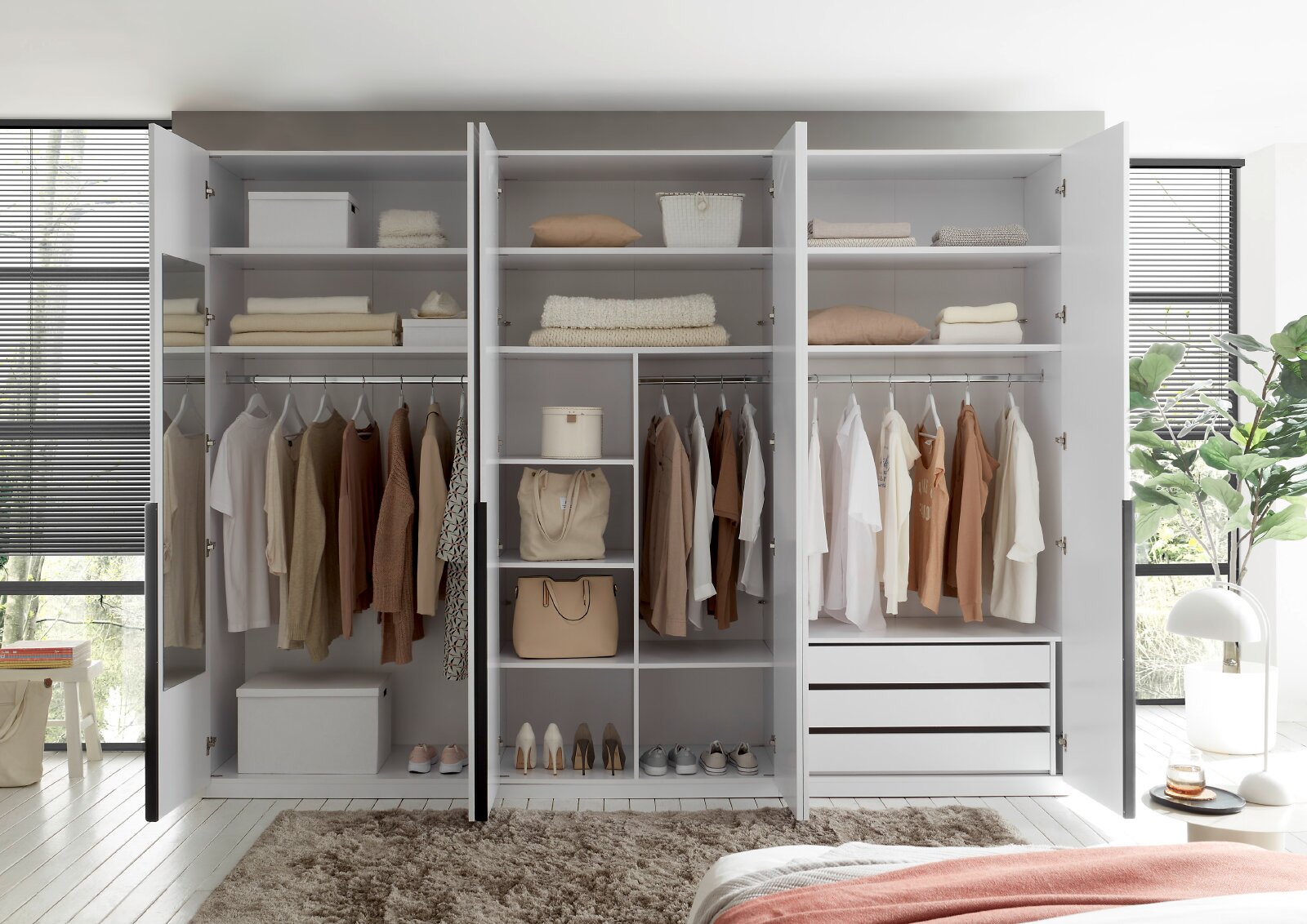 Großer weißer Kleiderschrank mit vier geöffneten Drehtüren, Kleiderstangen, Regalfächern und drei Schubladen, frontal in einem hellen Schlafzimmer gezeigt