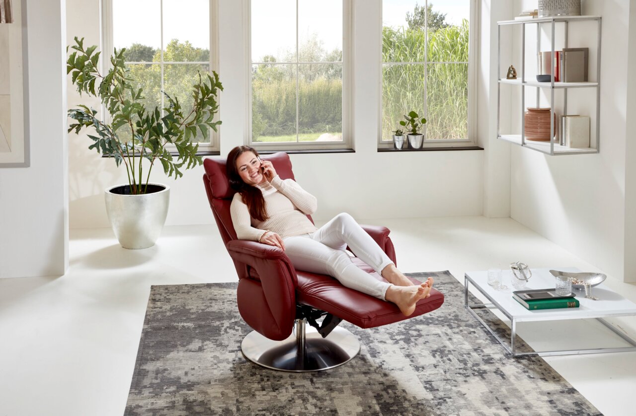 Een vrouw zit ontspannen in een rode tv-fauteuil in een lichte woonkamer met grote ramen, gezien vanuit een zijdelingse hoek.