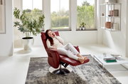 Een vrouw zit ontspannen in een rode tv-fauteuil in een lichte woonkamer met grote ramen, gezien vanuit een zijdelingse hoek.