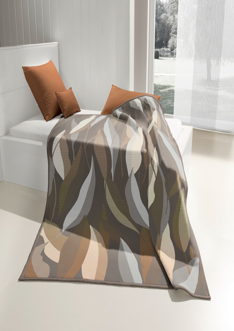 biederlack Woonkamerkleed COTTON HOME Een deken met een abstract bladmotief in aardetinten, gedrapeerd over een bed in een lichte kamer met uitzicht naar buiten.