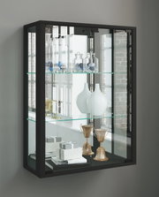 Schwarze Glasvitrine mit Glastüren und zwei Glasböden, bestückt mit Dekogläsern, weißen Vasen, Büchern und goldfarbenen Pokalen, fotografiert in leichter Frontansicht mit Perspektive von links.