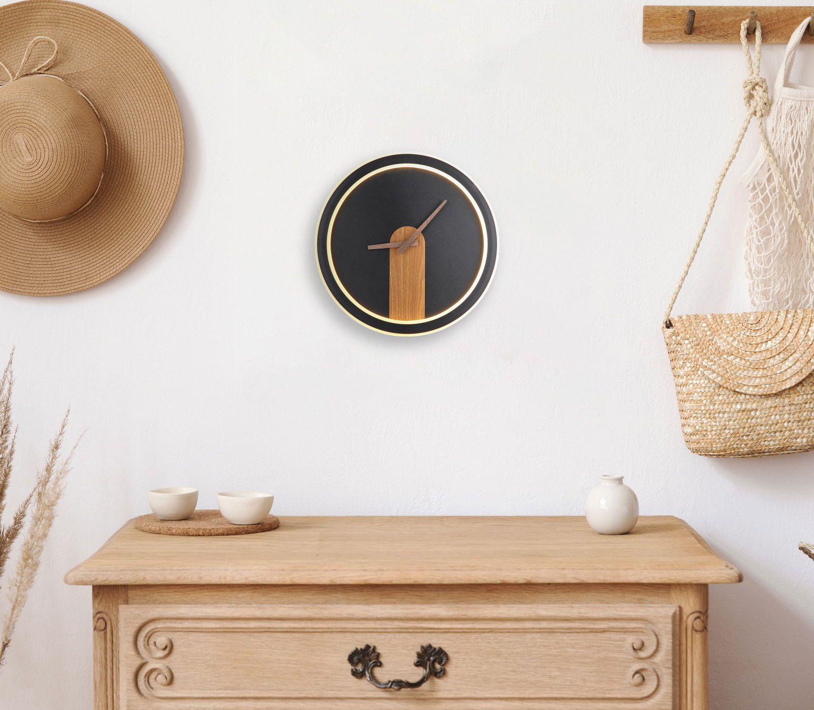 Moderne Wanduhr SUSSY mit schwarzem Zifferblatt und Holzdetails, frontal an einer weißen Wand über einer Kommode platziert.