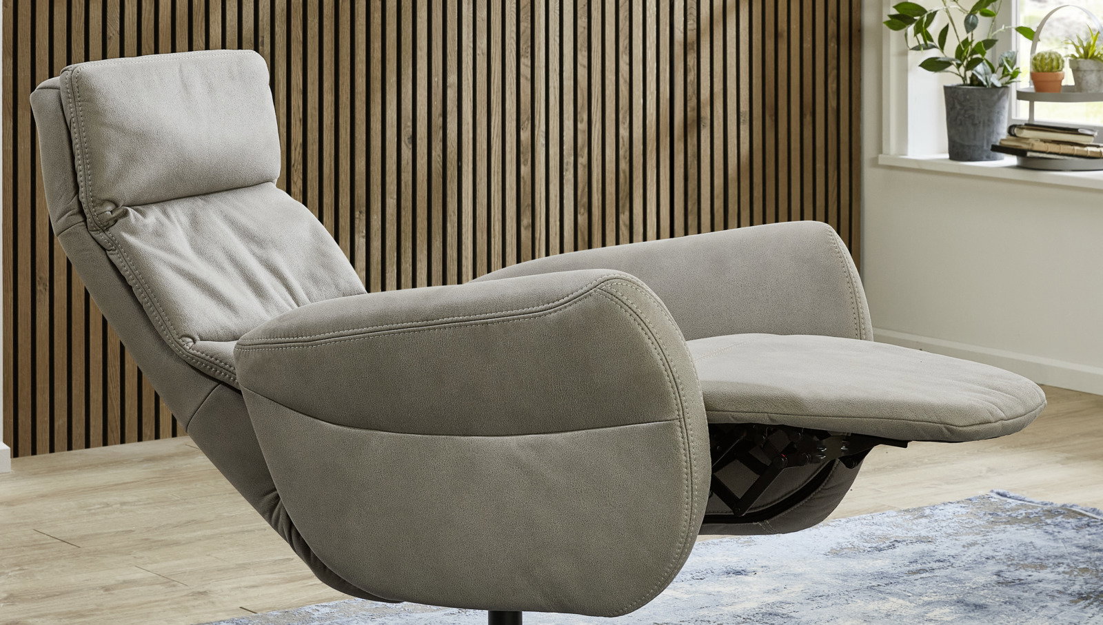 Grauer Relaxsessel in seitlicher Perspektive mit hochgeklappter Fußstütze vor einer Holzwand.