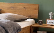Modernes Holzbett mit breitem Kopfteil und grauer Bettwäsche neben Nachttisch mit Uhr, Radio und kleiner Kaktus-Deko, seitliche Perspektive in Nahaufnahme