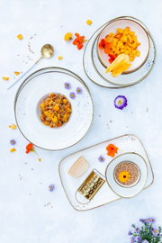 Draufsicht auf ein stilvolles Food-Arrangement mit weißem Geschirr: Schüssel mit gewürfelter Mango und essbaren Blüten, Teller mit herzhaftem Getreide-Gemüse-Mix, dazu Käse, goldene Reibe, Löffel und Körner auf hellem Hintergrund.