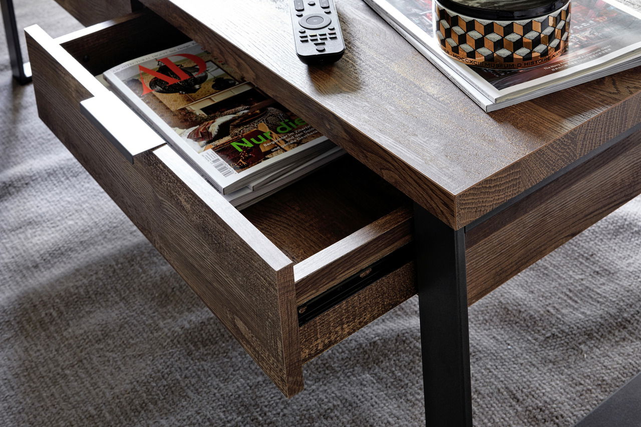 Nahaufnahme eines Couchtisches mit offener Schublade, in der Zeitschriften liegen. Auf der Tischplatte befinden sich eine Fernbedienung und weitere Zeitschriften. Perspektive von oben.