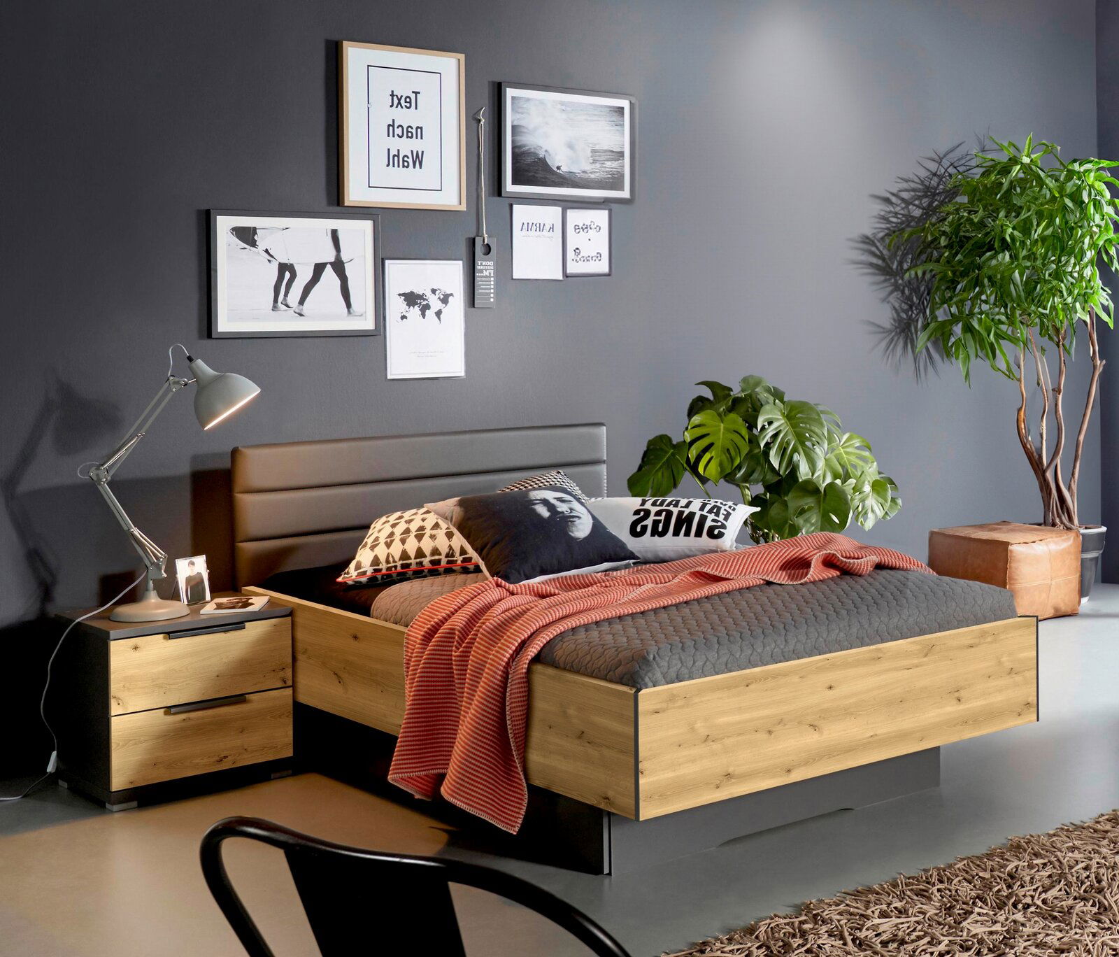 Modernes Holzbett mit gepolstertem grauen Kopfteil im Schlafzimmer, mit Nachttisch und Lampe, grauer Tagesdecke und roter Decke, Wandbildern und Zimmerpflanzen – Perspektive: schräg von vorne/seitlich.