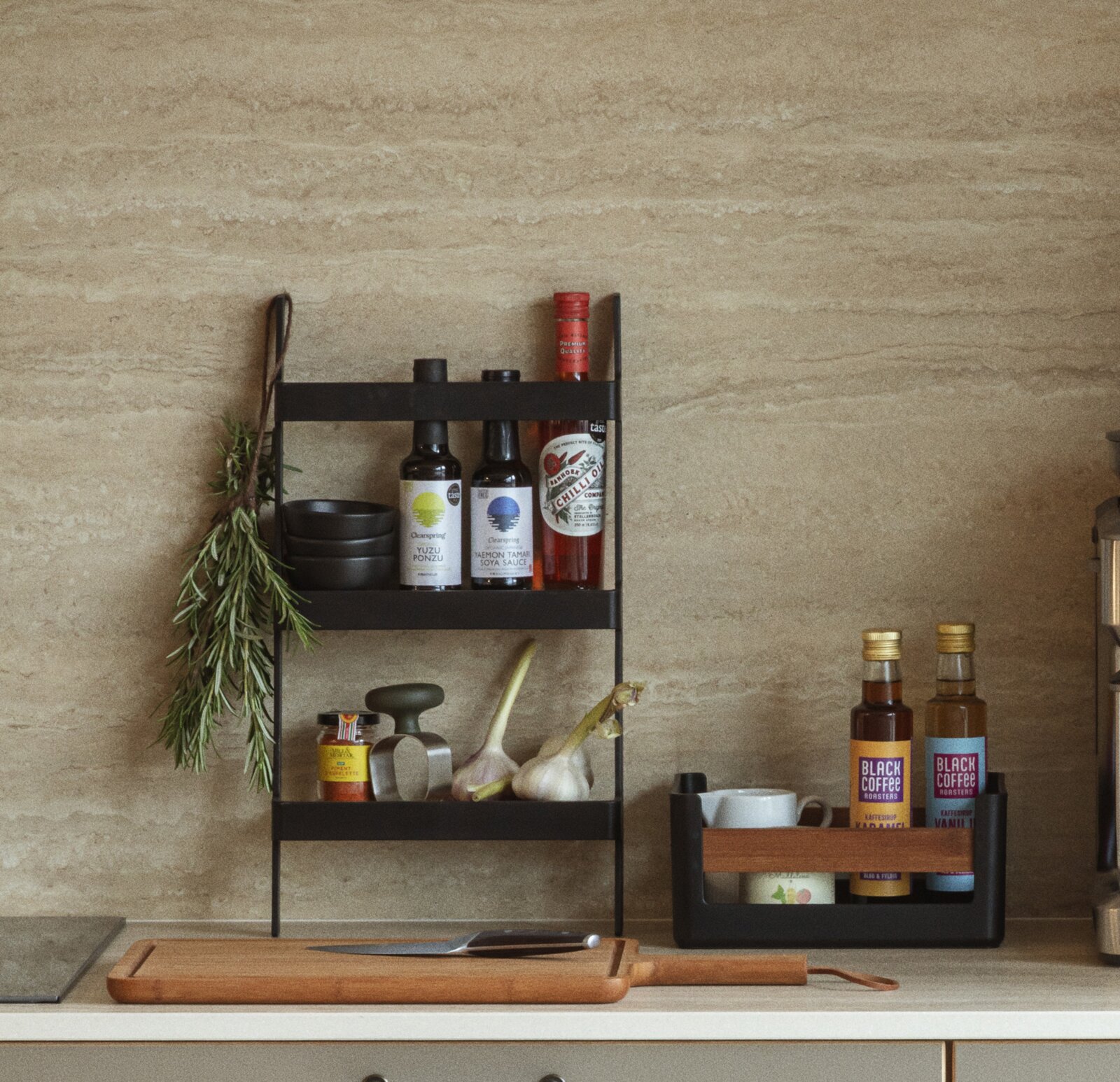 Eva Solo Lebensmittelkiste S NORDIC KITCHEN Schwarzes, dreistöckiges Küchenregal mit Flaschen, Schalen, Kräutern und Knoblauch auf einer Arbeitsplatte vor beiger Steinwand, frontale Perspektive.