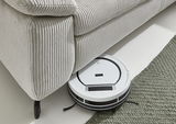 Ein Saugroboter reinigt den Boden unter einer hellen Polsterrundecke aus geripptem Stoff. Perspektive von oben.