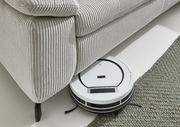 Ein Saugroboter reinigt den Boden unter einer hellen Polsterrundecke aus geripptem Stoff. Perspektive von oben.