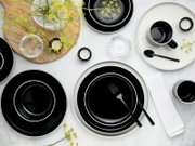 Draufsicht auf ein modern gedecktes Tisch-Arrangement mit schwarzem Geschirr mit weißem Rand, Tasse und Schalen, schwarzem Besteck, Teelichtern, Glas-Schälchen, Holzbrett und gelben Blüten auf hellem Hintergrund